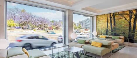 Una sala de estar moderna con ventanas amplias, vista a la ciudad y un mural de un bosque en la pared.