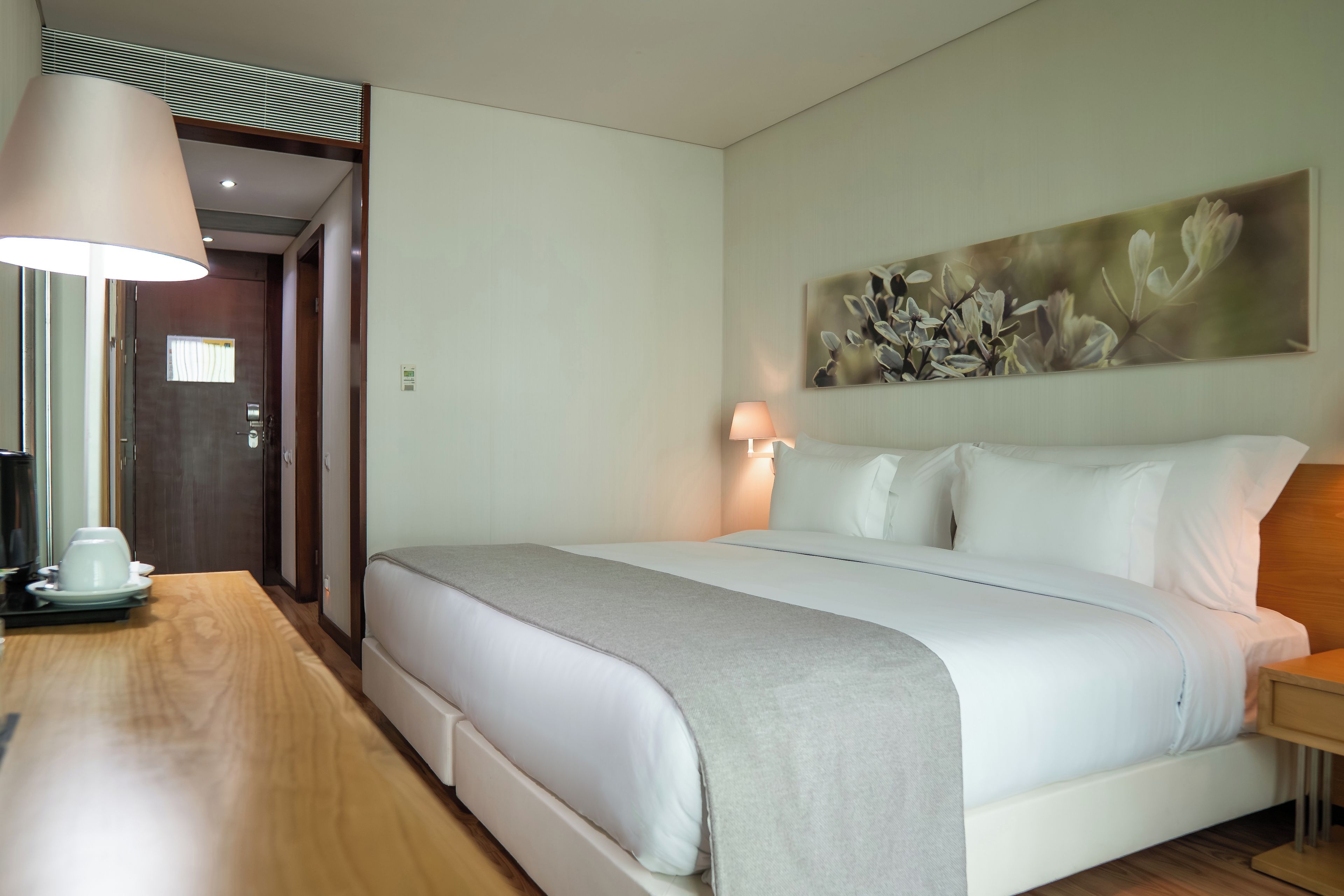 A hotel room with a bed, bedside table, lamp, and a painting on the wall.