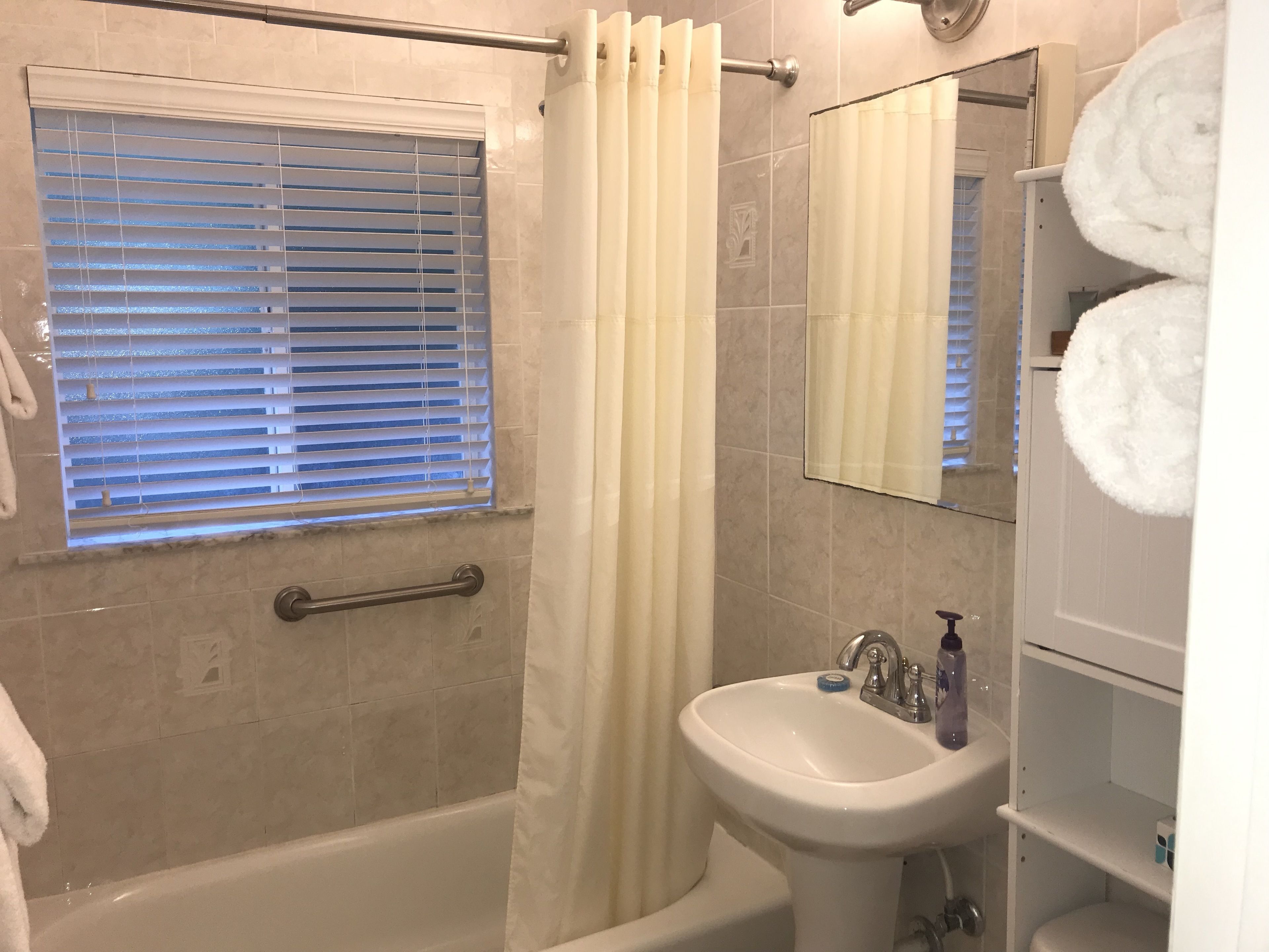 Un baño con bañera, ducha, lavamanos y ventana con persianas.