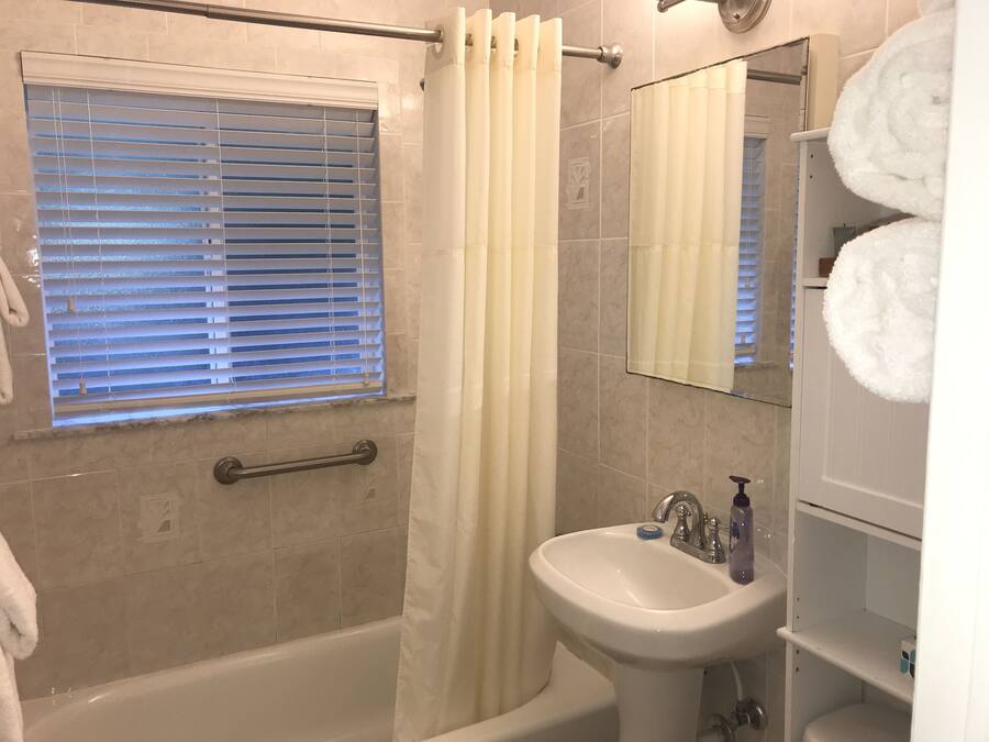 Un baño con bañera, ducha, lavamanos y ventana con persianas.