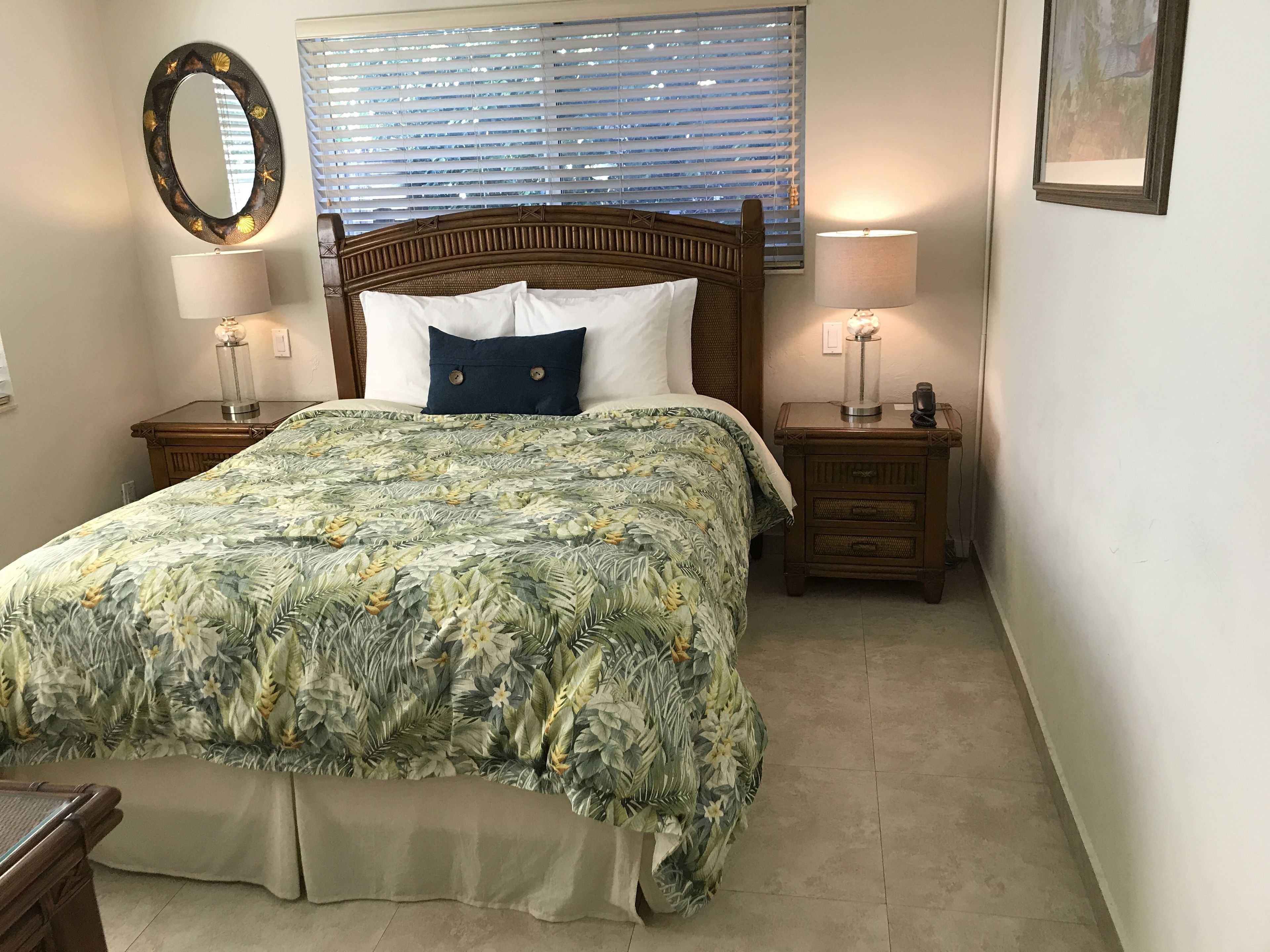A bedroom with a bed, two bedside tables, a nightstand, a mirror, and a window with blinds.