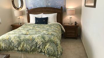 Une chambre à coucher comprenant un lit, deux tables de chevet, une commode, un miroir et une fenêtre avec des stores.