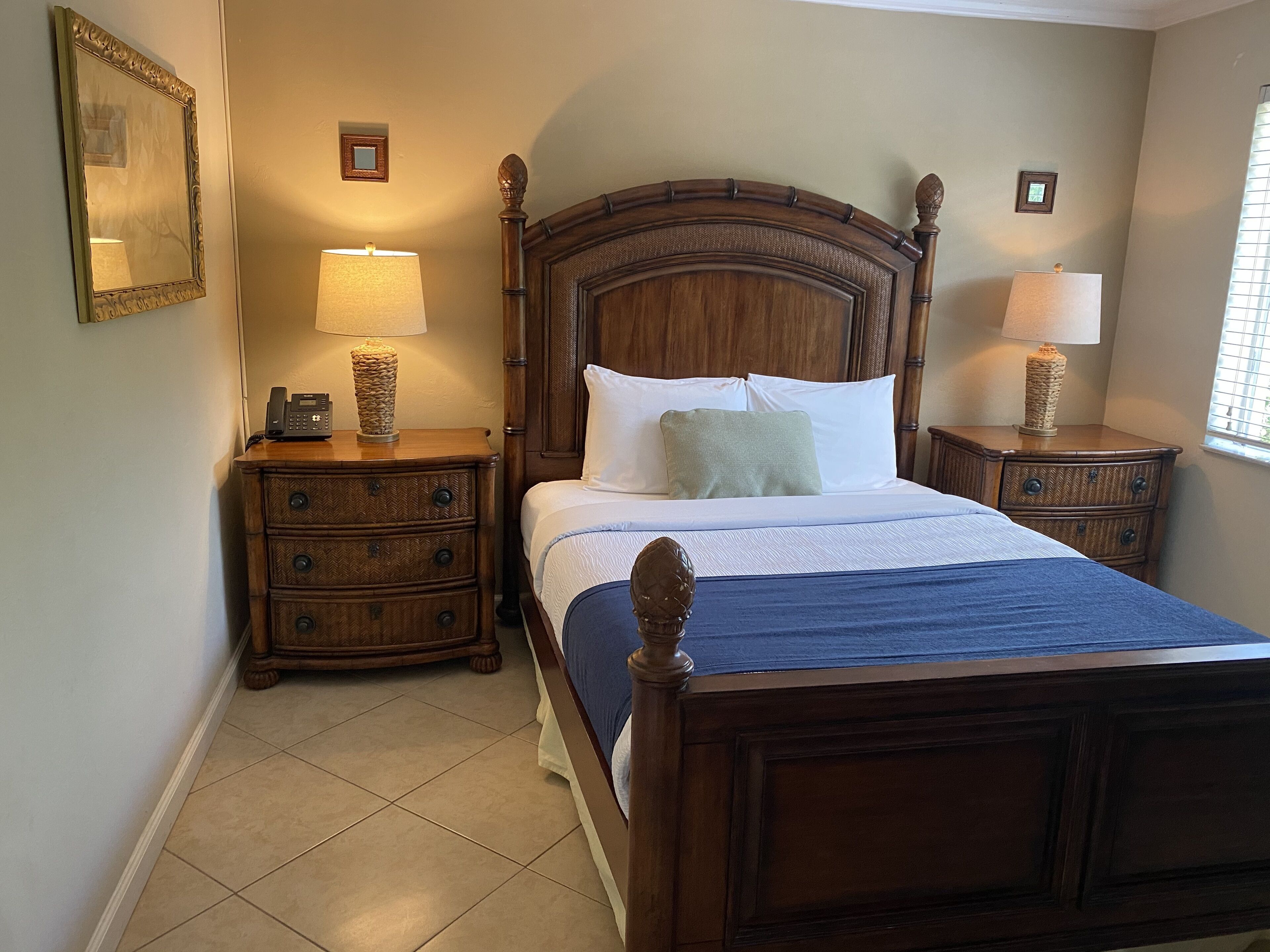 Quarto com cama de madeira, duas mesas de cabeceira com lumin árias, um espelho e uma pequena imagem na parede.