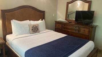 Una cama bien hecha con ropa de cama blanca y azul, un aparador de madera con espejo y televisión, y una mesita de noche con lámpara.