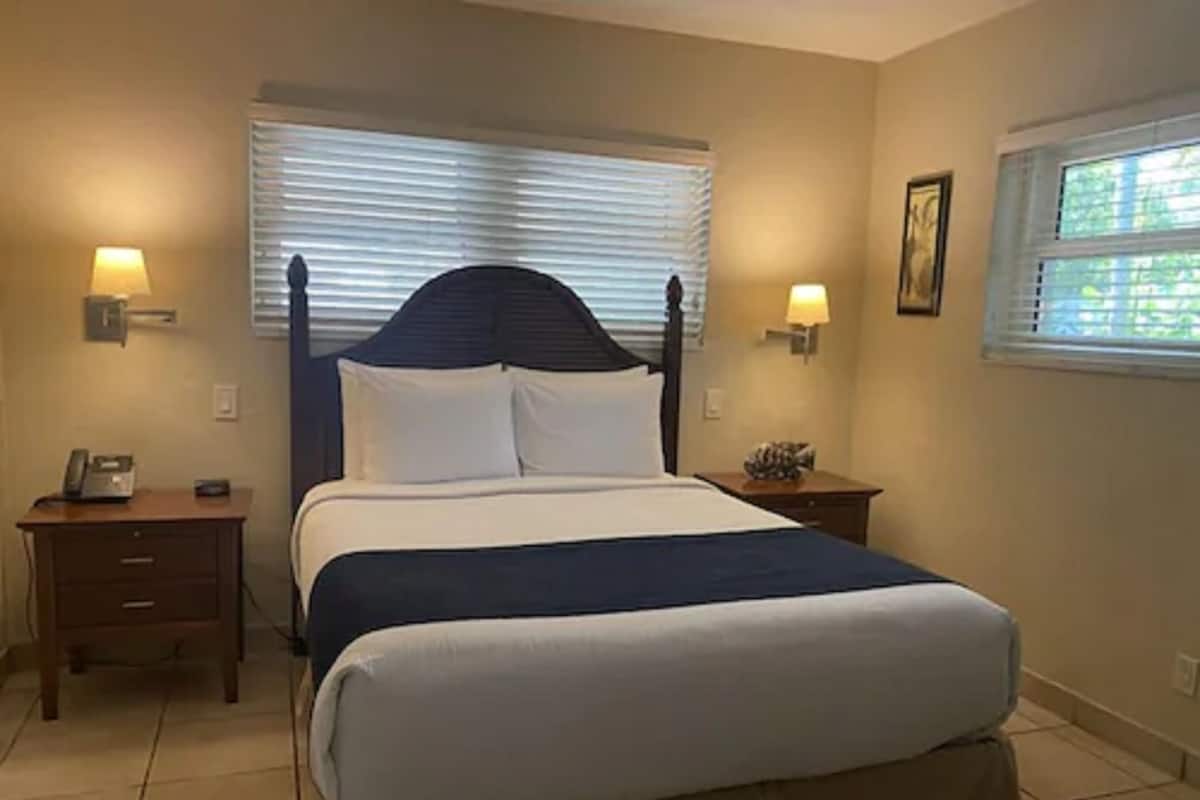 A neatly made bed with a dark headboard, two bedside tables with lamps, and a window with blinds.