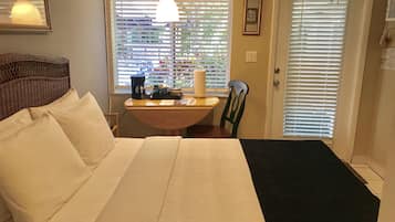 A bedroom with a bed, a table with a coffee maker, a chair, and a window with blinds.