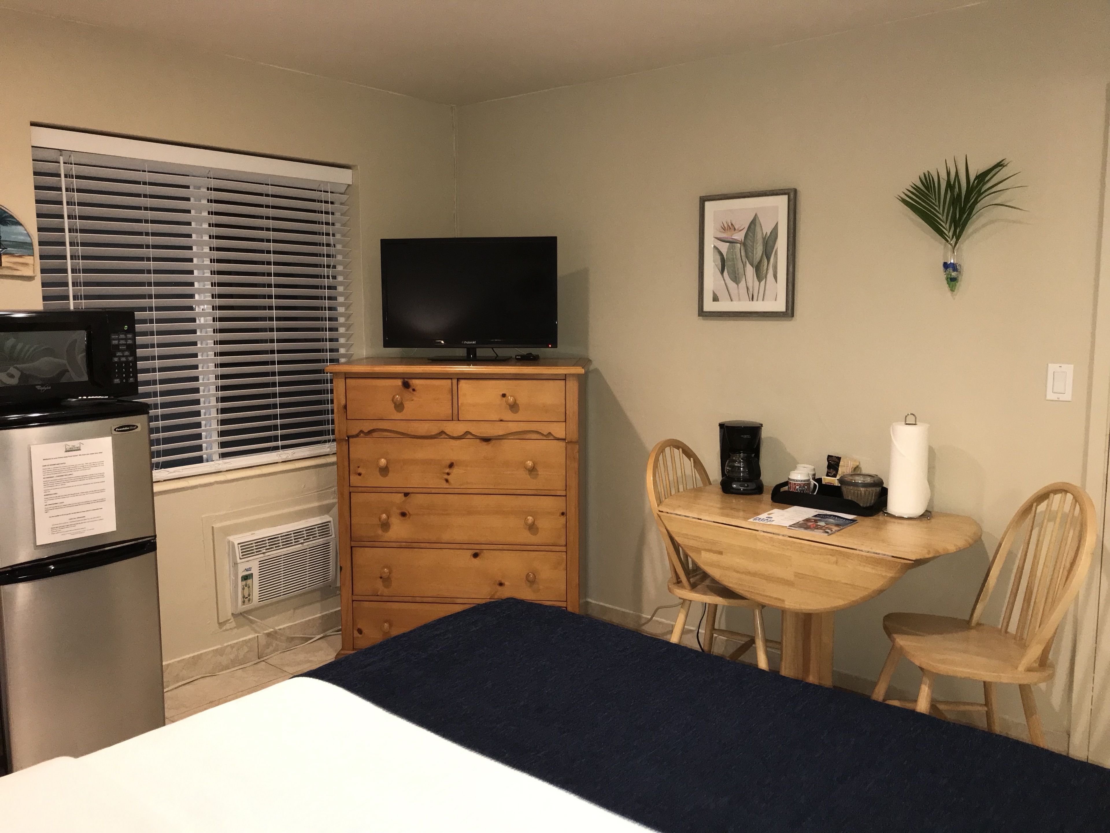 Habitación de hotel pequeña con cama, cómoda de madera, televisión, mesa de comedor con sillas y ventana con persianas.