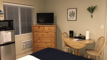 Habitación de hotel pequeña con cama, cómoda de madera, televisión, mesa de comedor con sillas y ventana con persianas.