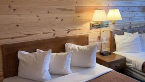 A hotel room with a wooden headboard, a bed with white bedding, two bedside lamps, and a brown textured rug. - Waterfront Inn - Mackinaw City (Mackinaw City)