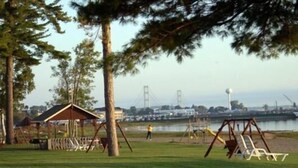 A park with a wooden play structure, a covered seating area, and a view of a bridge and water in the background. - Waterfront Inn - Mackinaw City (Mackinaw City)