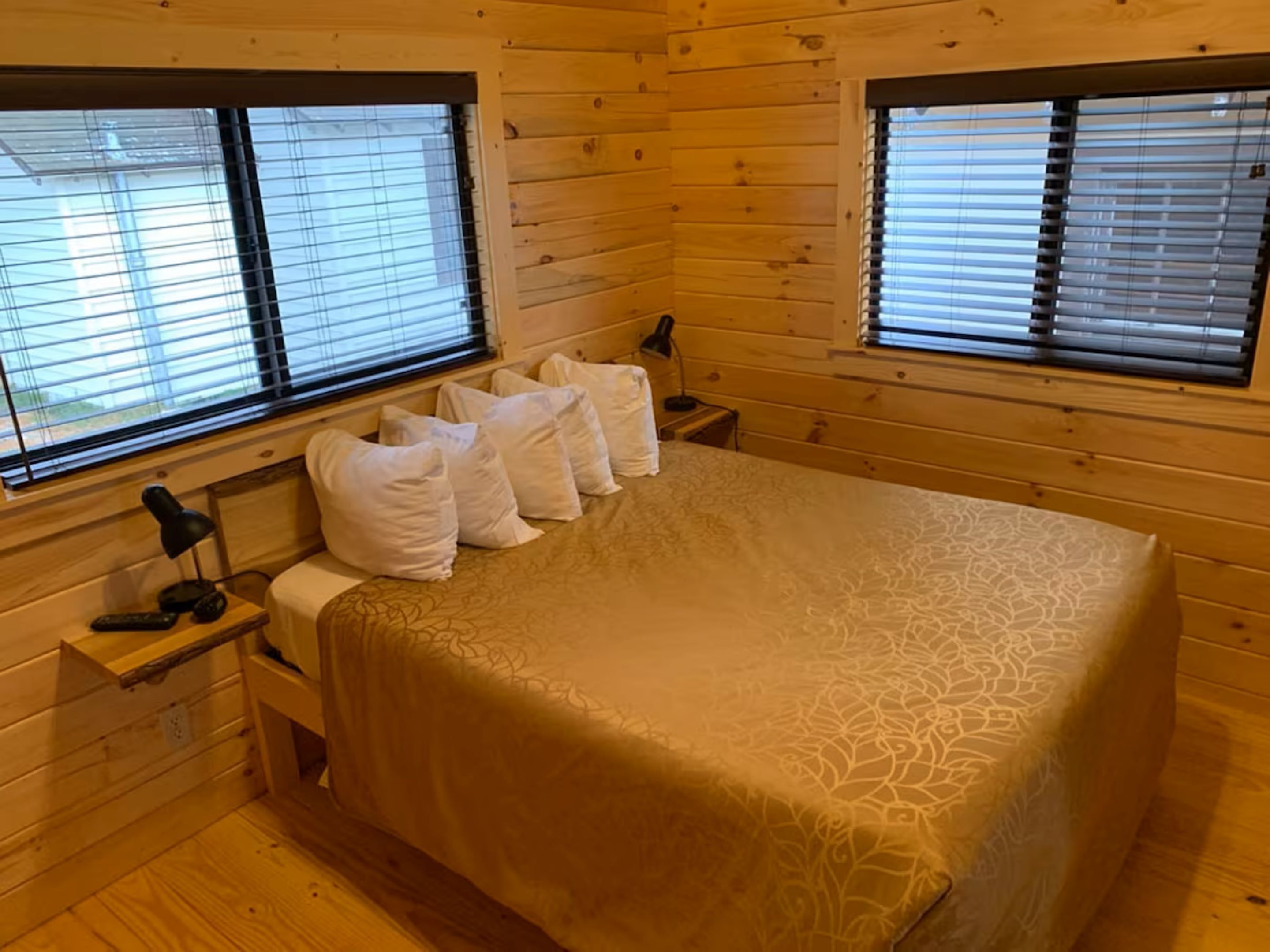 Una habitación de cabaña de madera con una cama, dos mesitas de noche y dos ventanas con persianas.