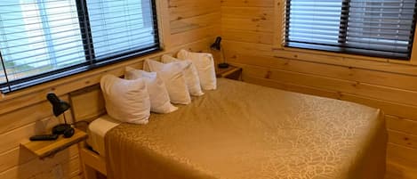 Una habitación de cabaña de madera con una cama, dos mesitas de noche y dos ventanas con persianas.
