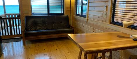 A cabin interior with a wooden floor, a sofa, a table, and windows with blinds.