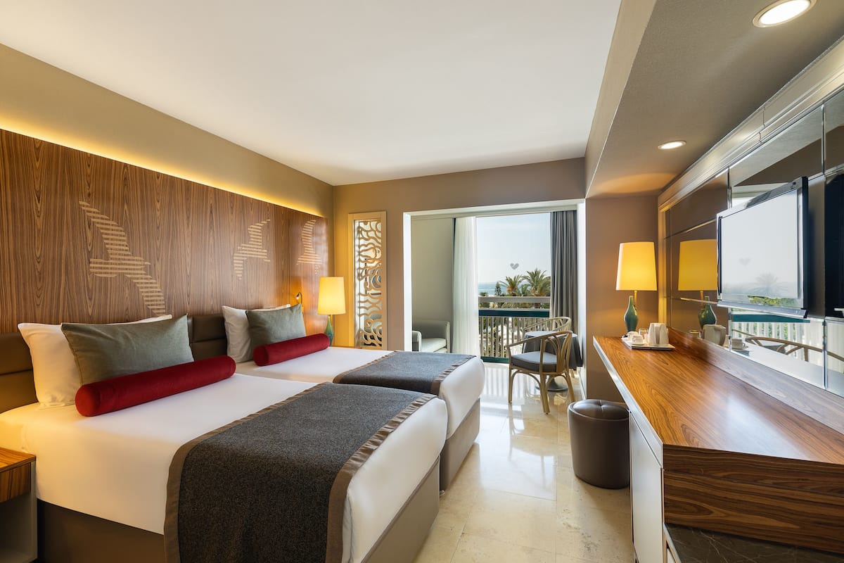 Quarto de hotel com duas camas, cabeceira de madeira, varanda com vista, escrivaninha de madeira e uma lixeira.