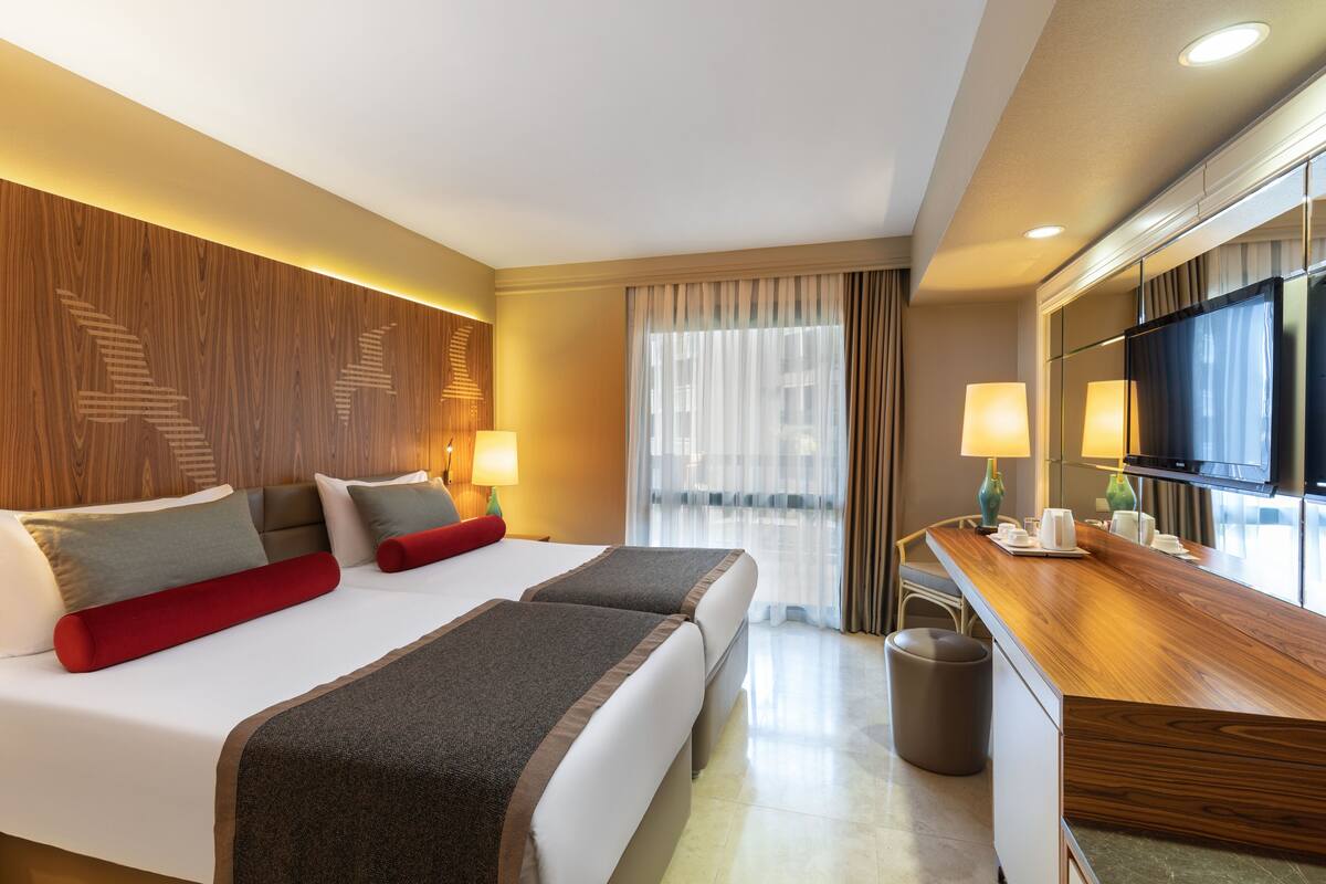 Quarto de hotel com uma cama grande, cabeceira de madeira, escrivaninha com televisão e janela com cortinas.
