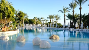Zona de piscina con sillones de descanso y sombrillas, rodeada de palmeras.