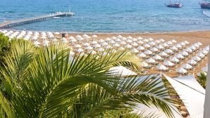 Una playa con hileras de sombrillas blancas y sillas reclinables, un muelle que se adentra en el mar y dos embarcaciones al fondo.