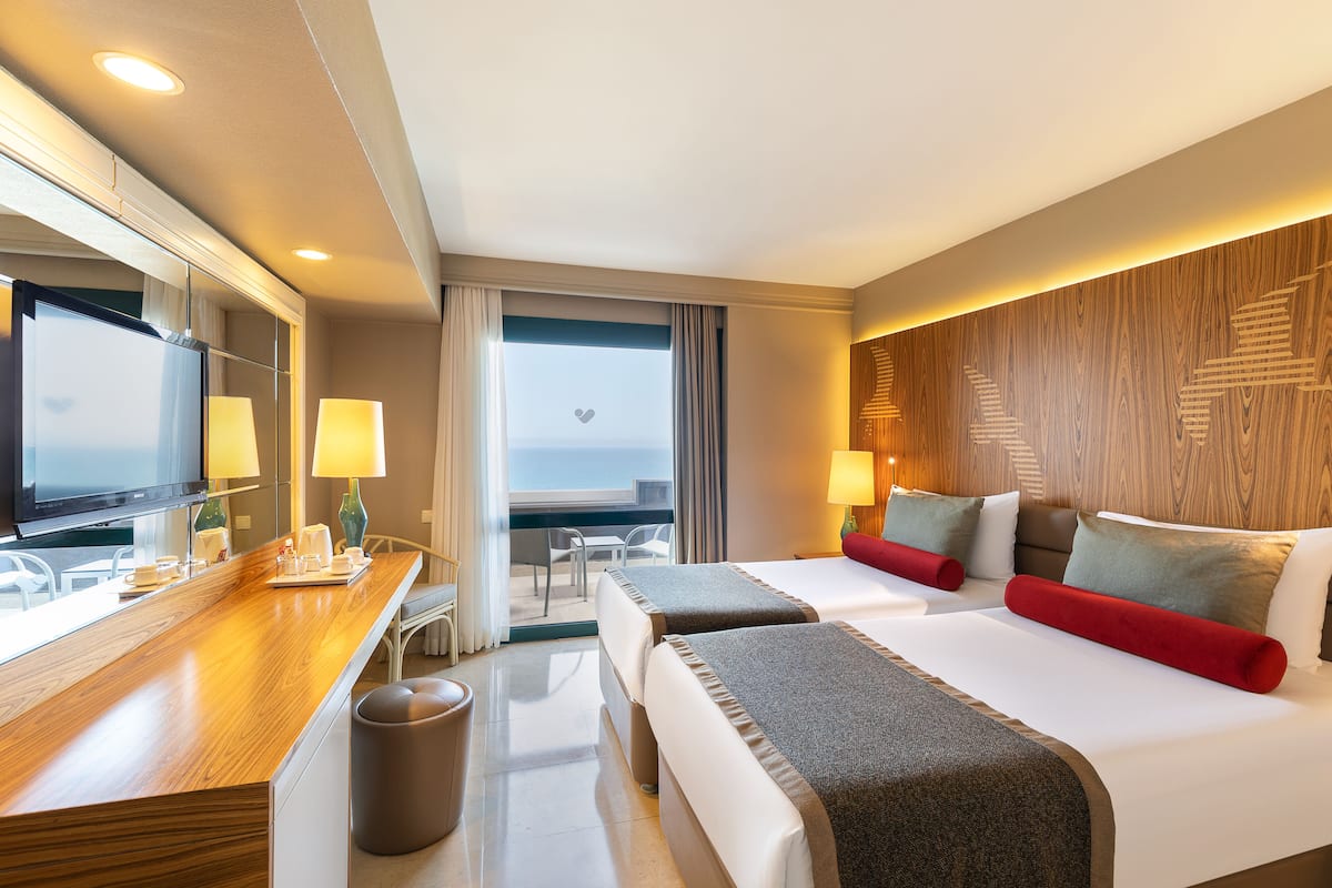 Quarto de hotel com escrivaninha de madeira, duas camas, televisão e varanda com vista.