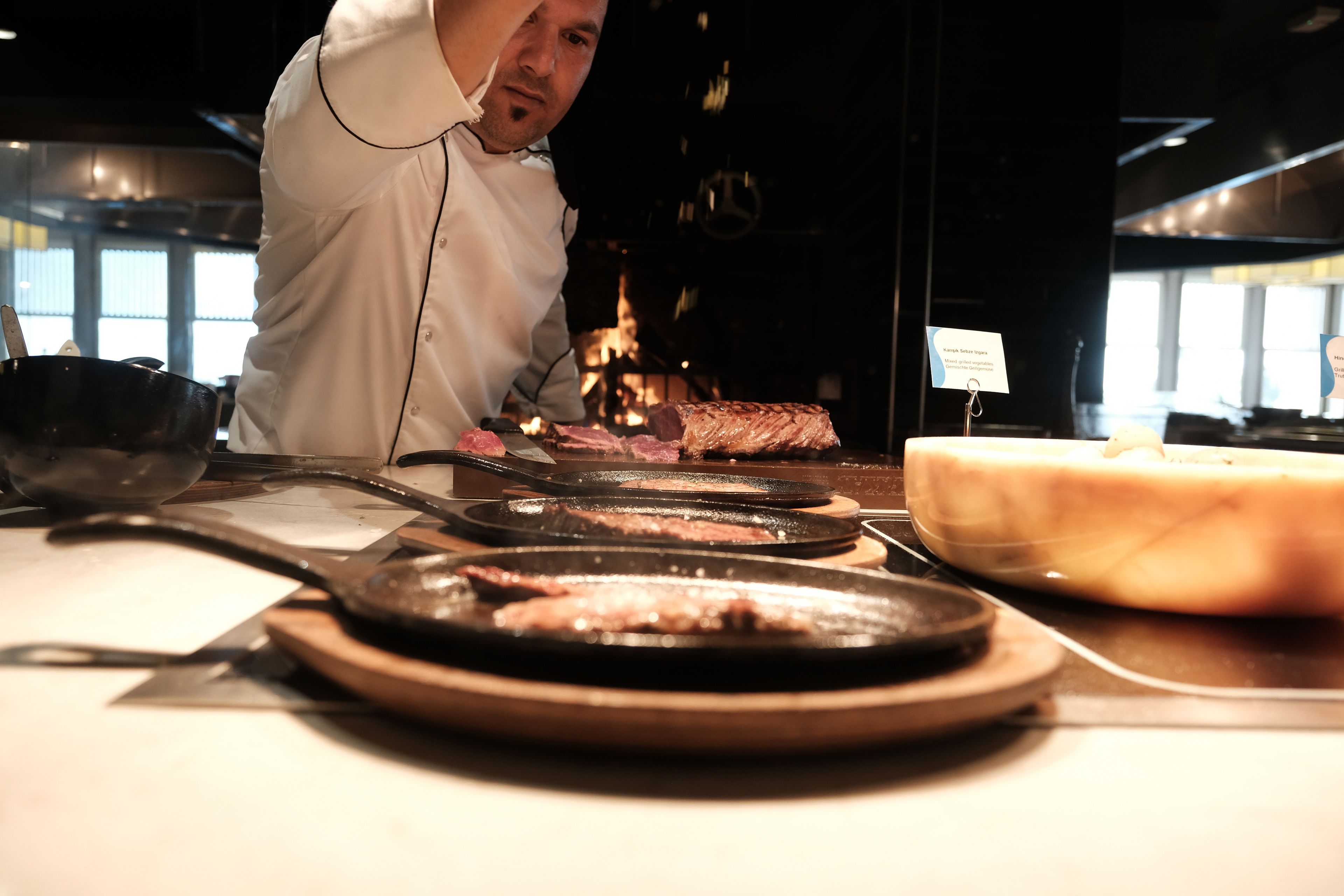 Un chef preparando comida en una cocina con una tabla de cortar grande, una sartén y una parrilla.