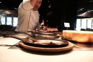 Un chef preparando comida en una cocina con una tabla de cortar grande, una sartén y una parrilla.
