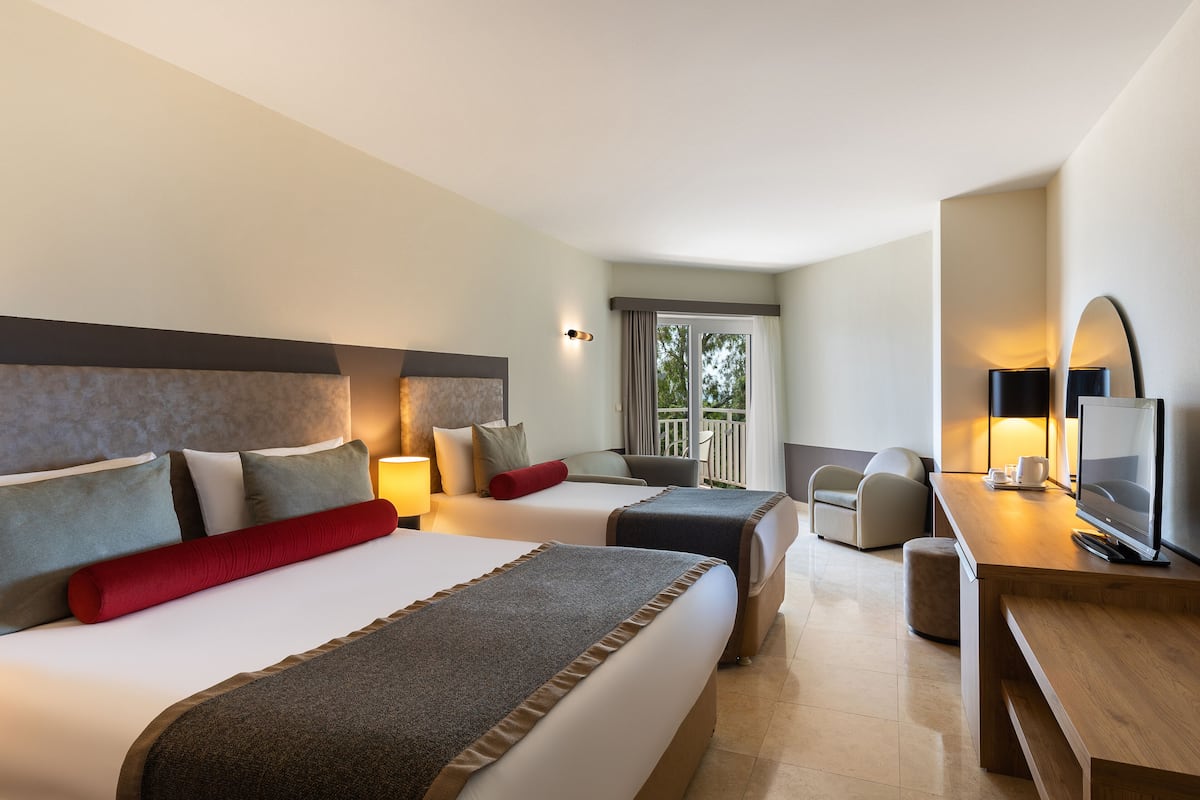 Quarto de hotel com duas camas, uma escrivaninha com televisão e varanda com vista.