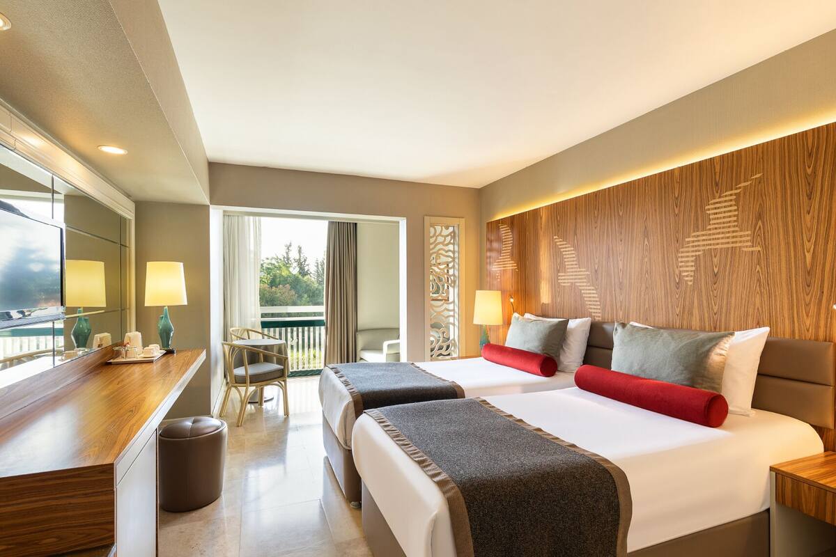 Quarto de hotel com duas camas, uma escrivaninha, uma cadeira e uma varanda.