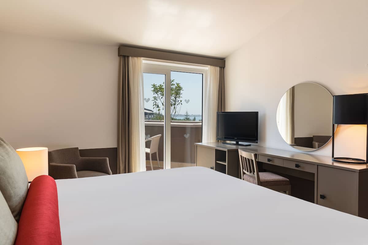 Quarto de hotel com cama, escrivaninha com televisão, cadeira, abajur e uma grande janela com vista para o exterior.