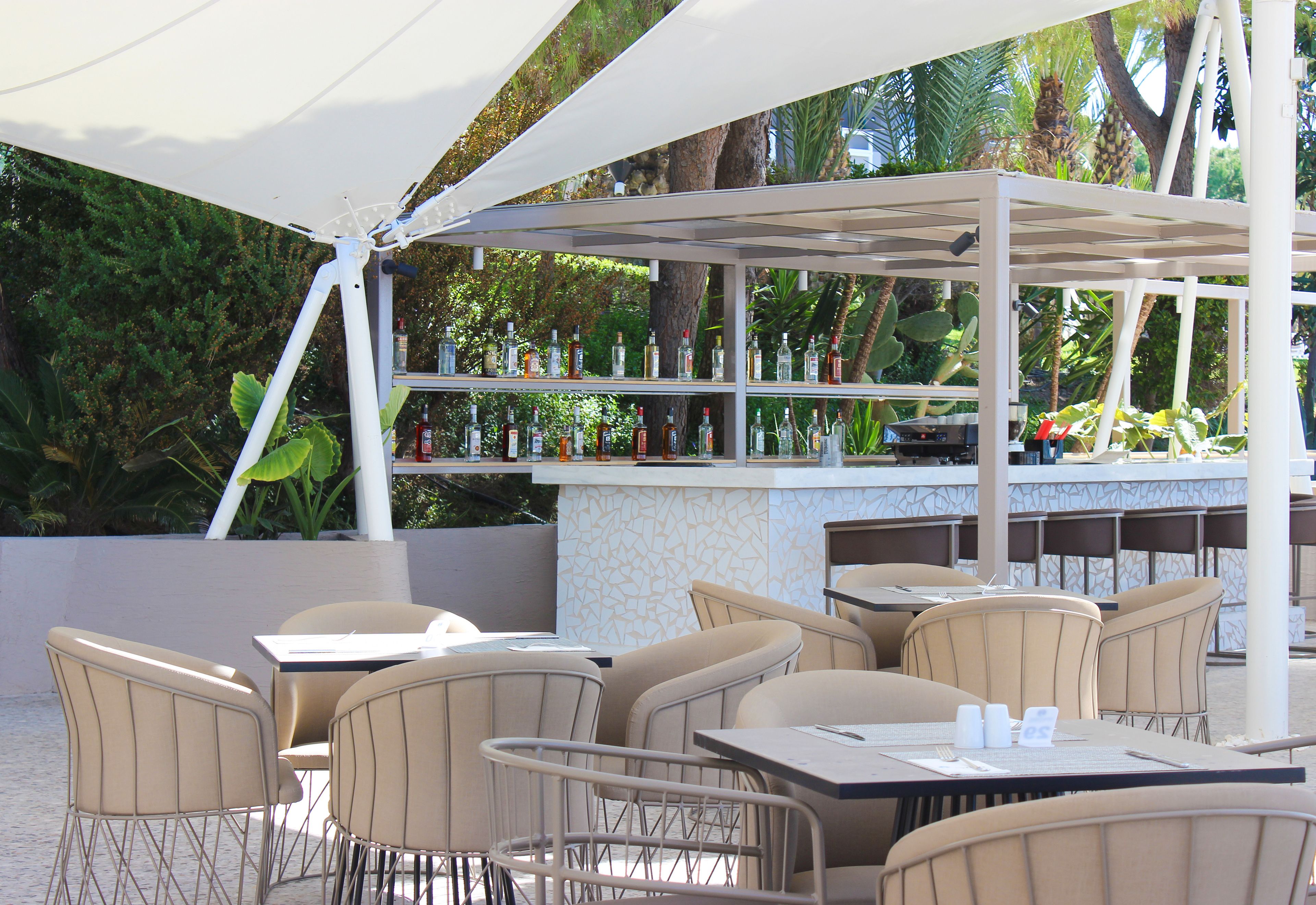 Un patio al aire libre con mesas y sillas, una zona de bar con botellas, y un toldo blanco.