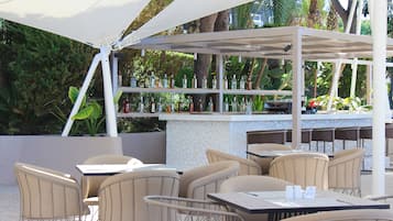 Een terras buiten met tafels en stoelen, een bar met flessen en een witte luifel.