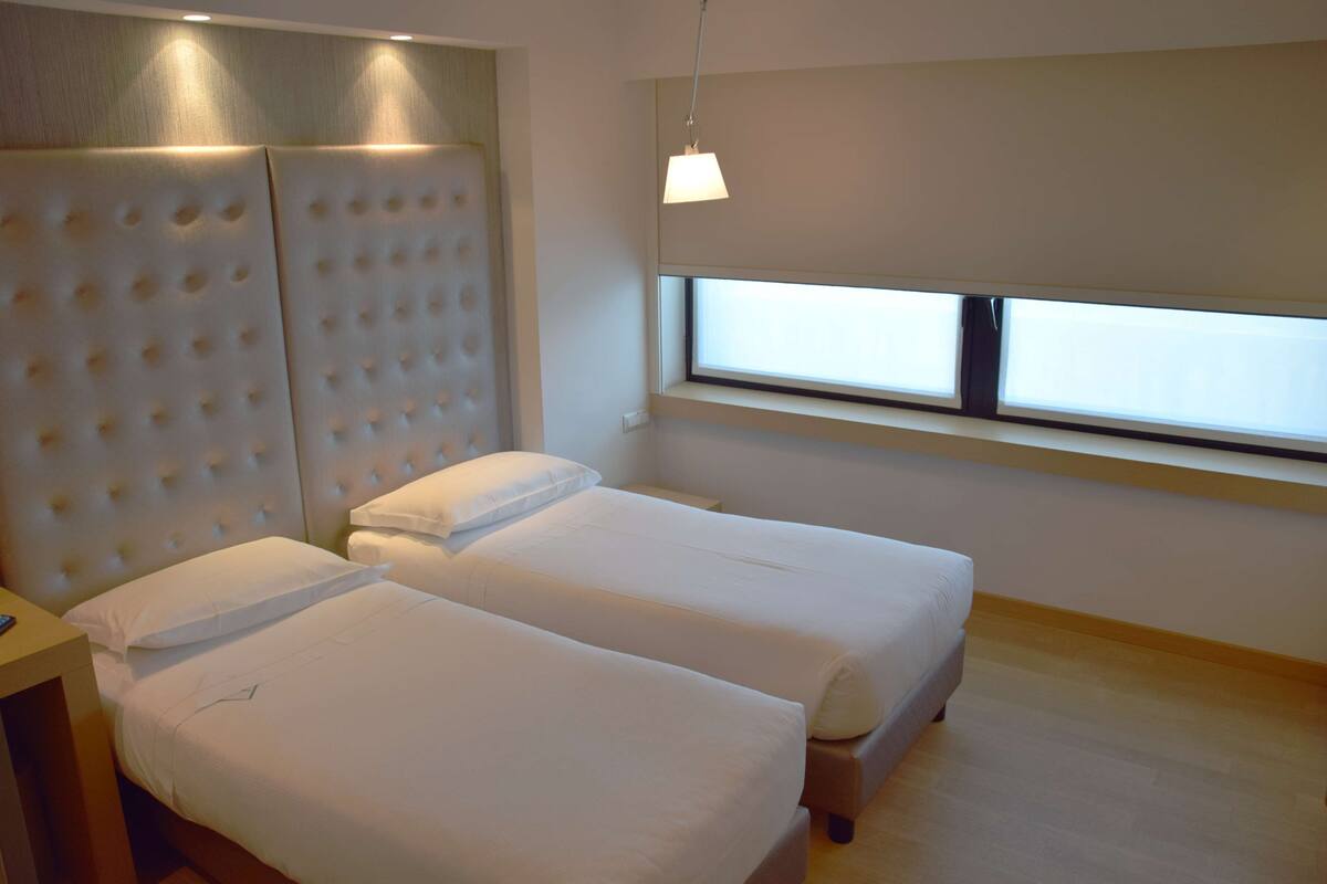 Een hotelkamer met twee enkele bedden, een groot gestoffeerd hoofdbord, een raam met luxaflex en een hanglamp.