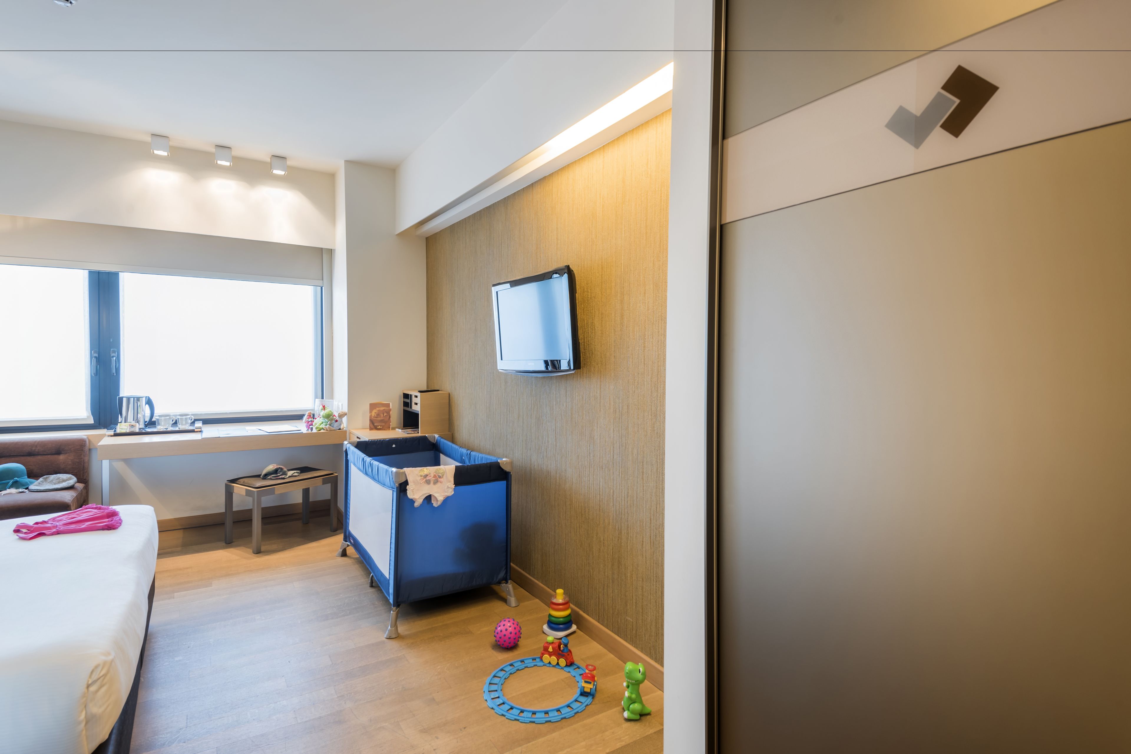 Ein Hotelzimmer mit einem Bett, einem Schreibtisch, einem Fernseher, einem blauen Reisebett und Spielzeug auf dem Boden.