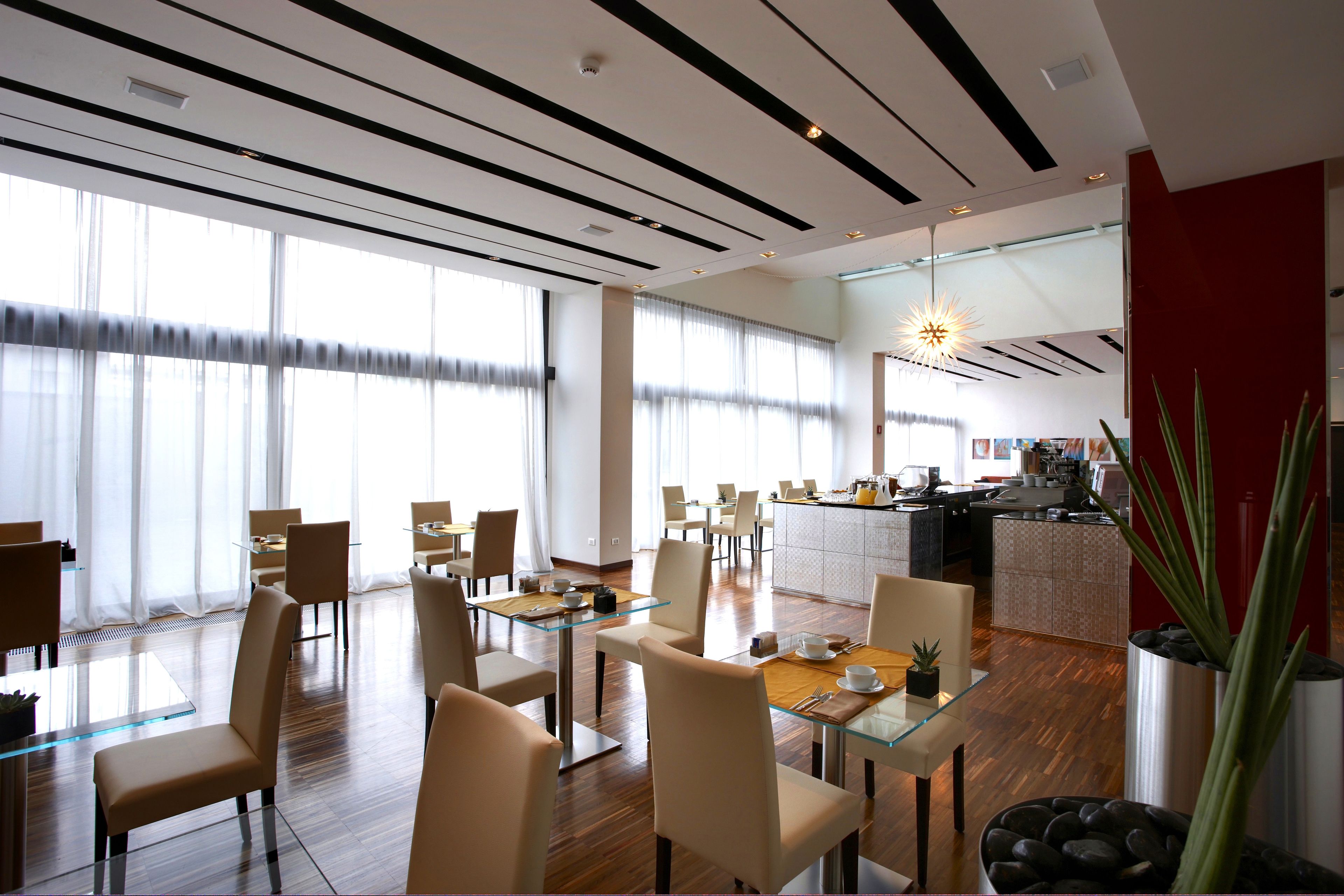 A modern dining area with glass tables and chairs, a kitchen in the background, and large windows with sheer curtains.
