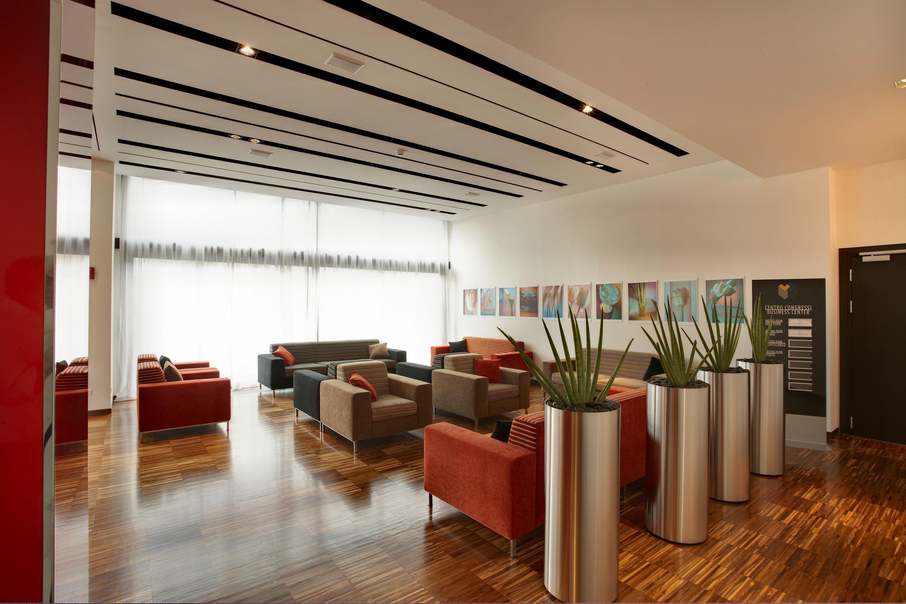 A modern lobby with wooden flooring, red and grey sofas, and tall planters.