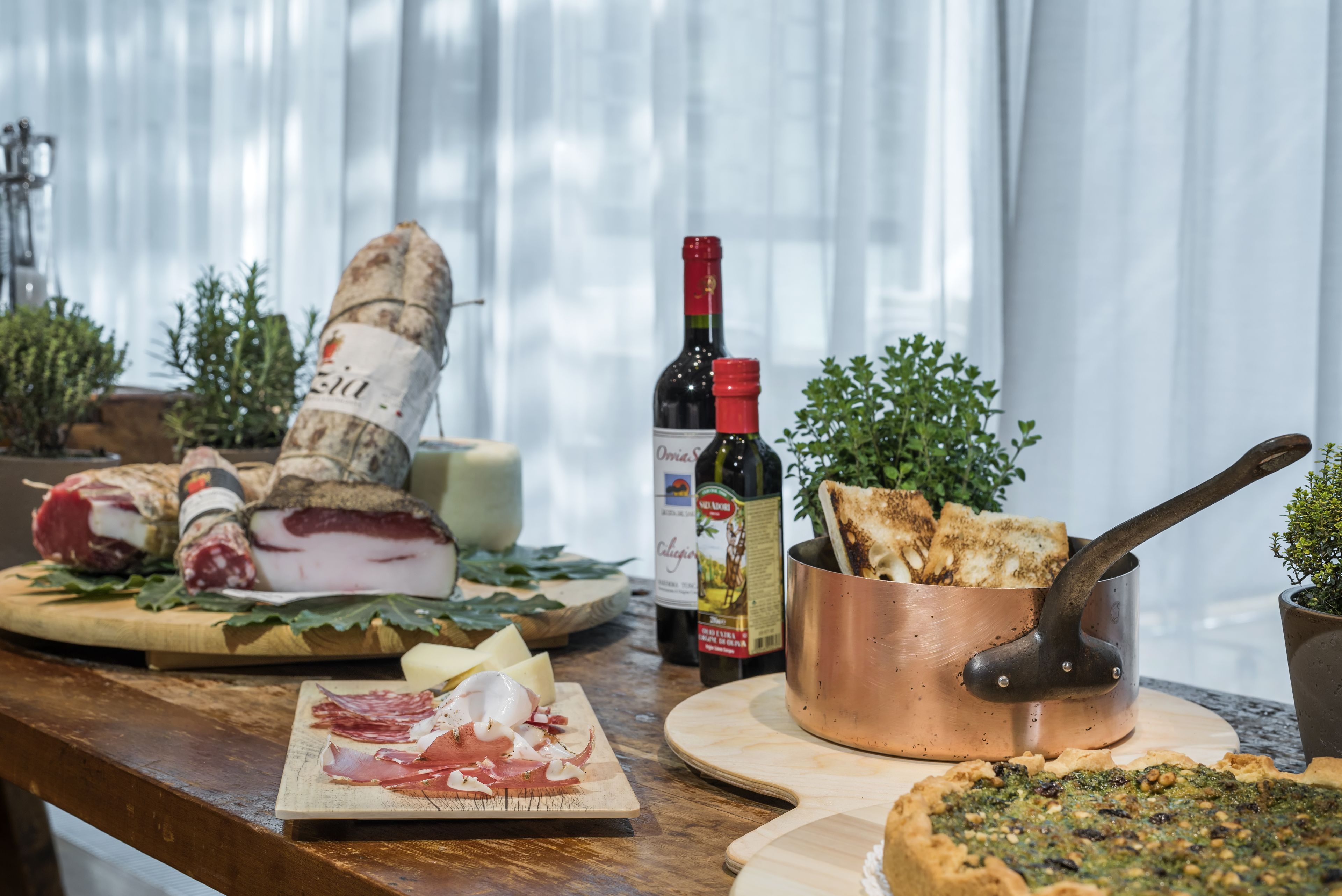 Ein Holztisch mit verschiedenen Speisen, darunter Wurstwaren, Käse und Pizza. Eine Weinflasche und ein Kupfertopf mit Brot sind ebenfalls vorhanden.