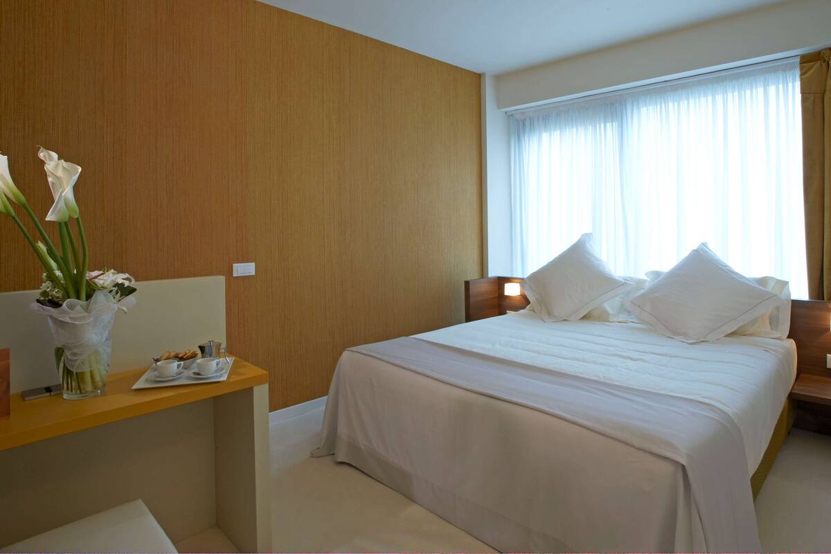Een hotelkamer met een bed, nachtkastjes, een bureau, een bloevas en een raam met gordijnen.