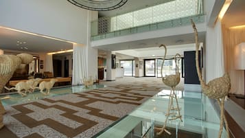 A spacious lobby with a high ceiling, a glass floor, and a patterned carpet. There are decorative sheep sculptures and a large, circular chandelier.