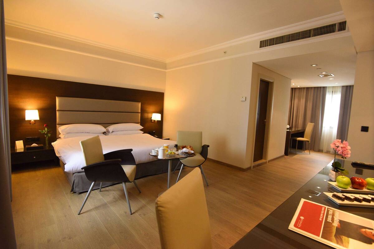Habitación de hotel con una cama grande, un escritorio con una silla, una pequeña mesa con comida y una lámpara en la mesita de noche.