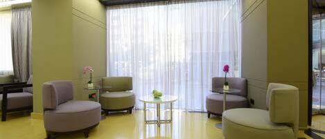 A modern hotel lobby with a yellow floor, two round tables with chairs, and a large window with sheer curtains.