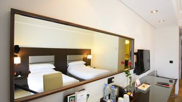 A hotel room with a large mirror reflecting a neatly made bed, a bedside table with a lamp, a TV, and a tray with toiletries.