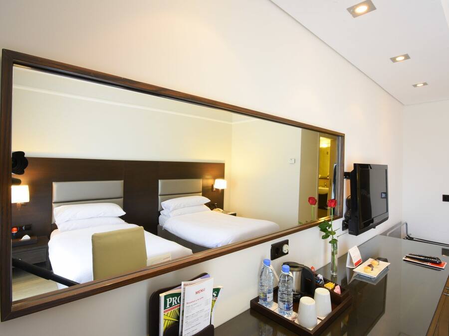 Habitación de hotel con un espejo grande que refleja una cama bien tendida, una mesita de noche con lámpara, un televisor y una bandeja con artículos de aseo.