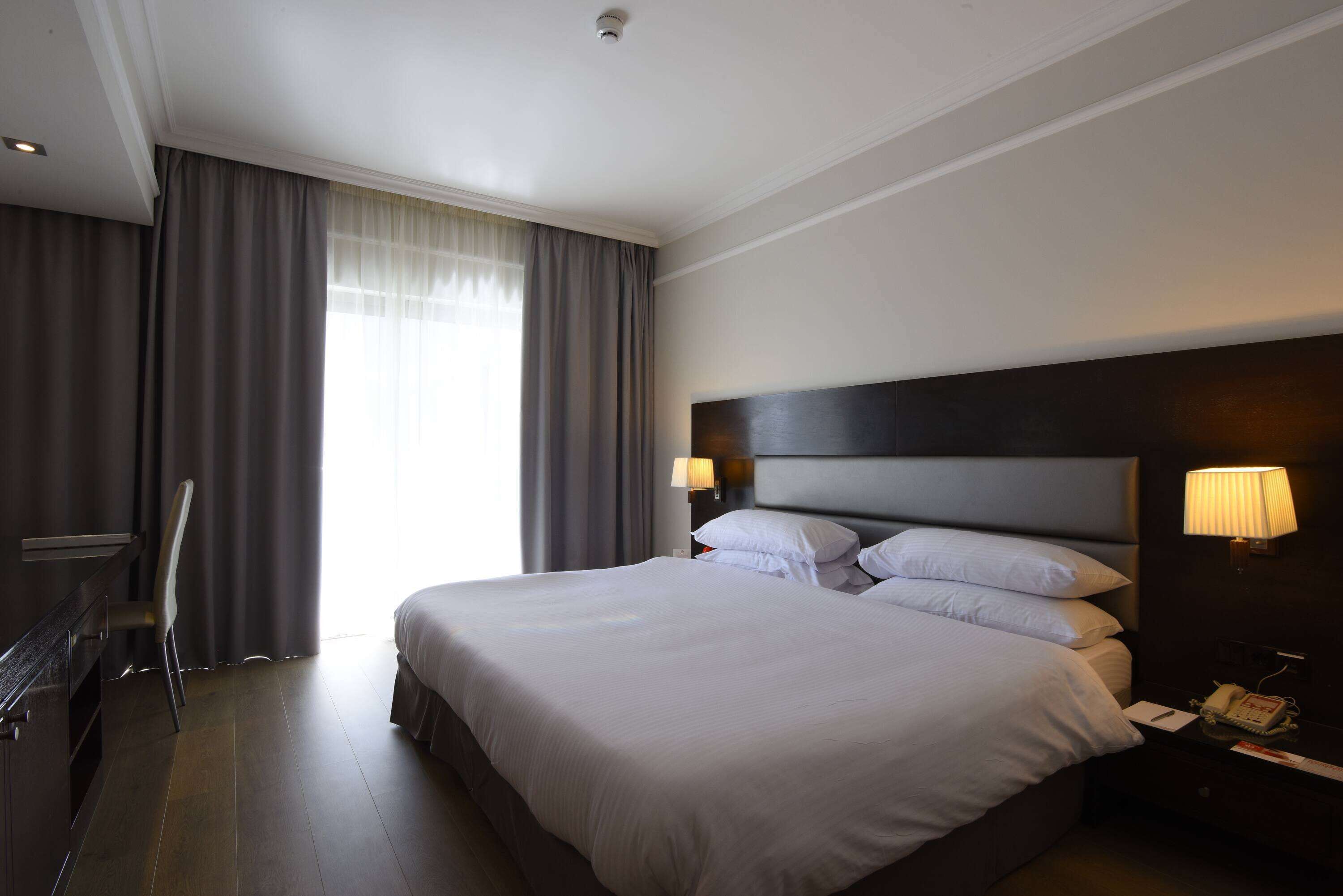 A hotel room with a large bed, a desk with a chair, a nightstand with a lamp, and a window with curtains.