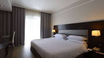 A hotel room with a large bed, a desk with a chair, a nightstand with a lamp, and a window with curtains.
