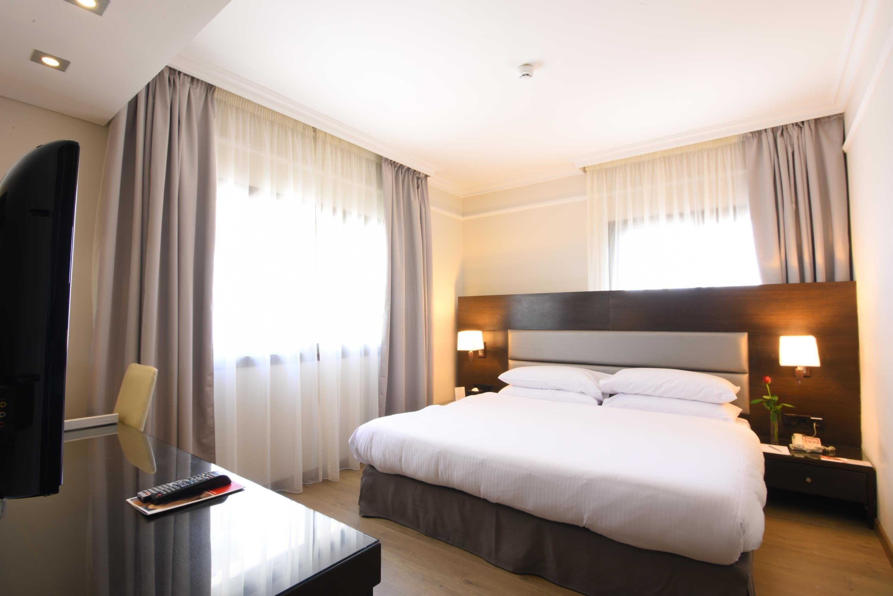 A hotel room with a large bed, bedside lamps, a desk with a remote control, and curtains.