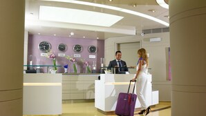 Un área de recepción con diseño moderno, que incluye un mostrador de recepción, un reloj que muestra la hora en varias ciudades y un arreglo floral decorativo.
