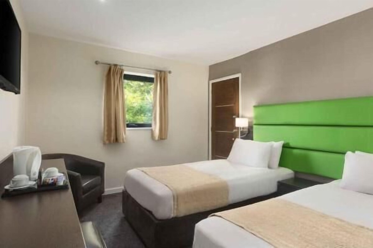 A hotel room with two beds, a desk, a chair, a window with curtains, and a green headboard.