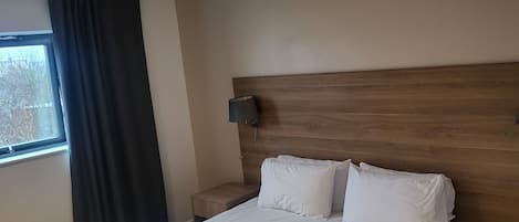 A hotel room with a large bed, a nightstand, a wall-mounted lamp, and a window with curtains.