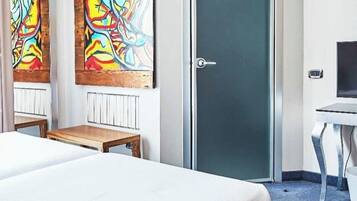 A hotel room with a bed, a desk, two abstract paintings, a door, and a radiator.
