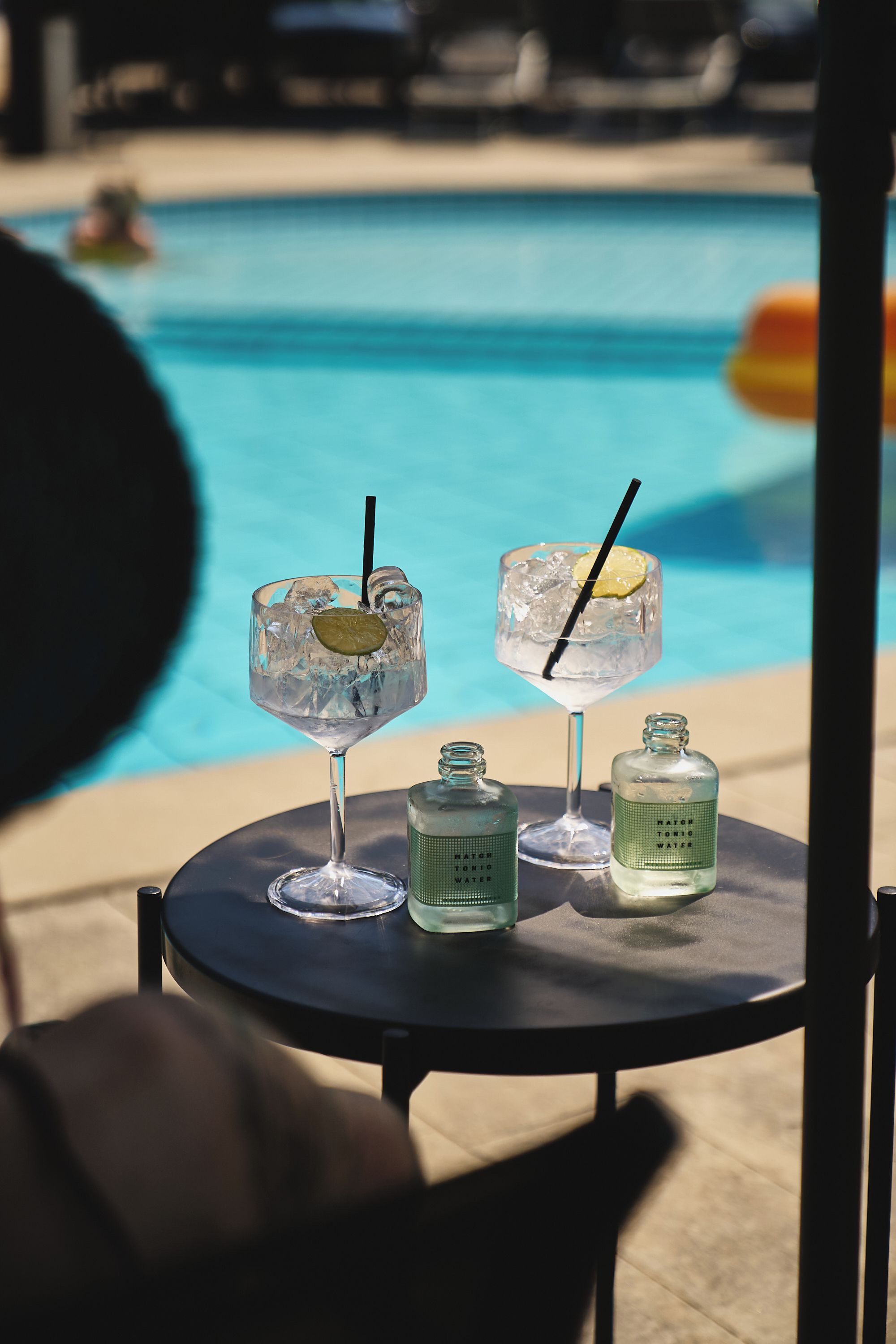 A table with two drinks, each with a lime wedge and straw, and two bottles labeled 'MATCH TONIC WATER' on a poolside setting.