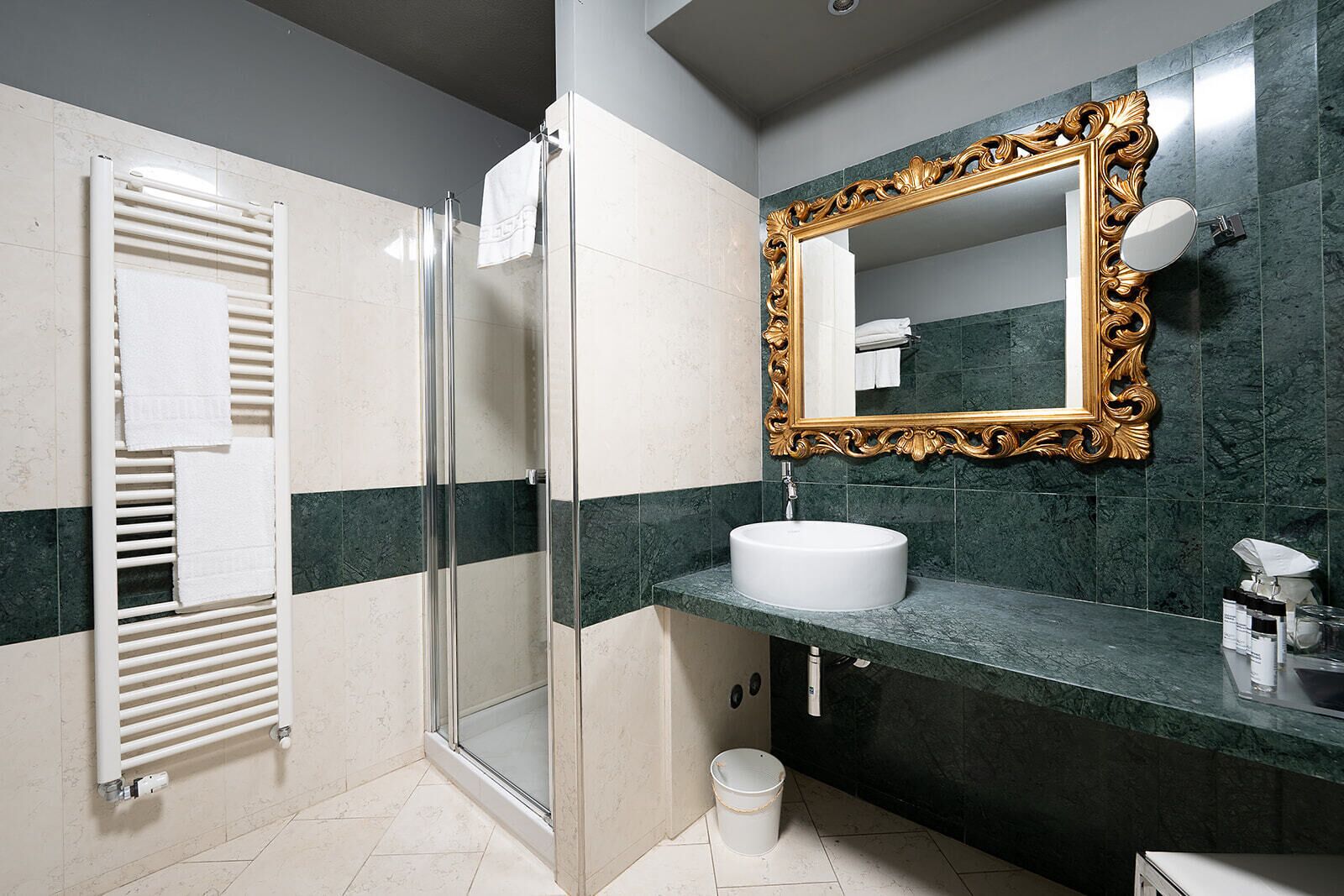 Un baño con cabina de ducha de vidrio, un lavamanos blanco, un espejo con marco dorado y un cubo blanco en el suelo.