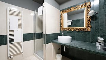 Un baño con cabina de ducha de vidrio, un lavamanos blanco, un espejo con marco dorado y un cubo blanco en el suelo.